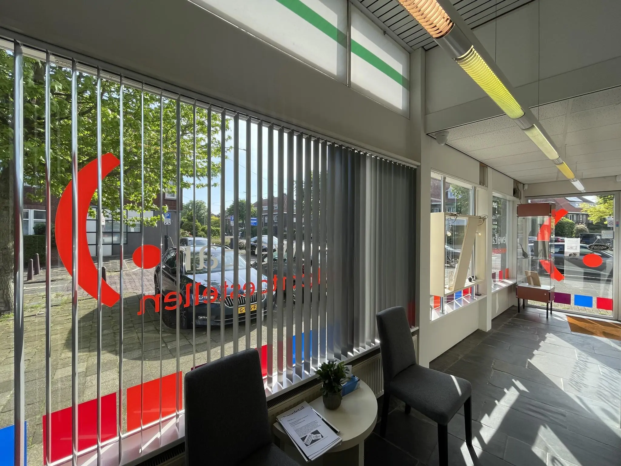 Winkelpand aan de Prinses Mariannelaan met grote ramen, verticale jaloezieën en kleurrijke raamstickers.